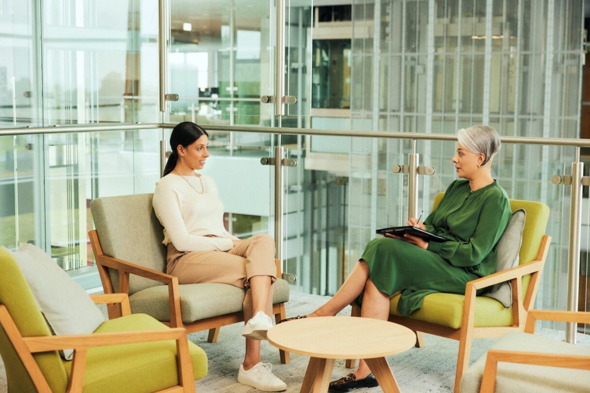 Duas mulheres jovens em reunião numa sala de escritório moderna, sentadas frente a frente com computadores portáteis e documentos, a discutir ideias de trabalho com expressão focada e colaborativa.