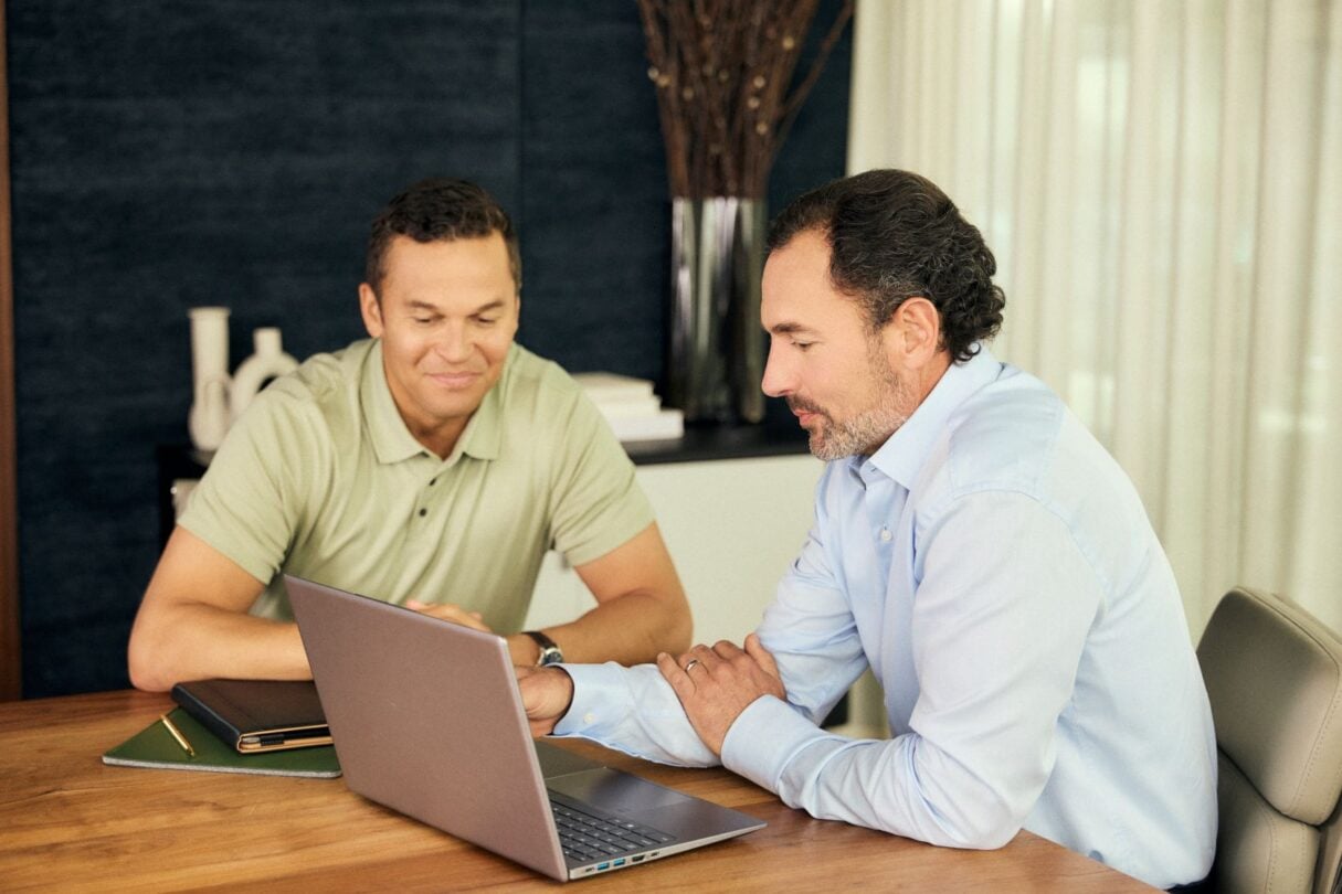 Dois jovens homens sentados lado a lado num escritório moderno, concentrados a trabalhar num computador portátil.