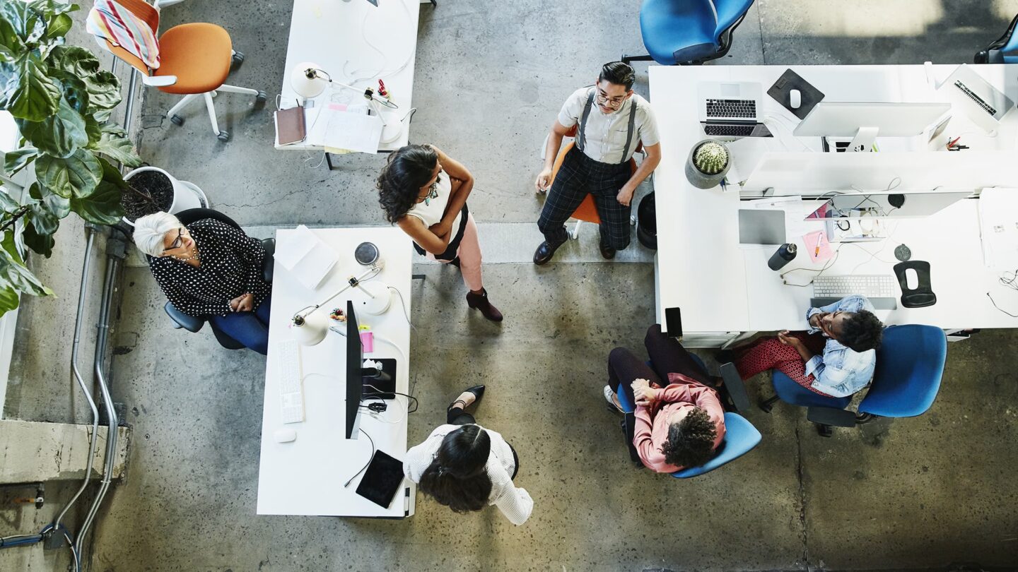 Vários colegas de trabalho estão reunidos numa oficina moderna. Estão sentados e de pé à volta de mesas com computadores portáteis, a conversar e a colaborar em tarefas.