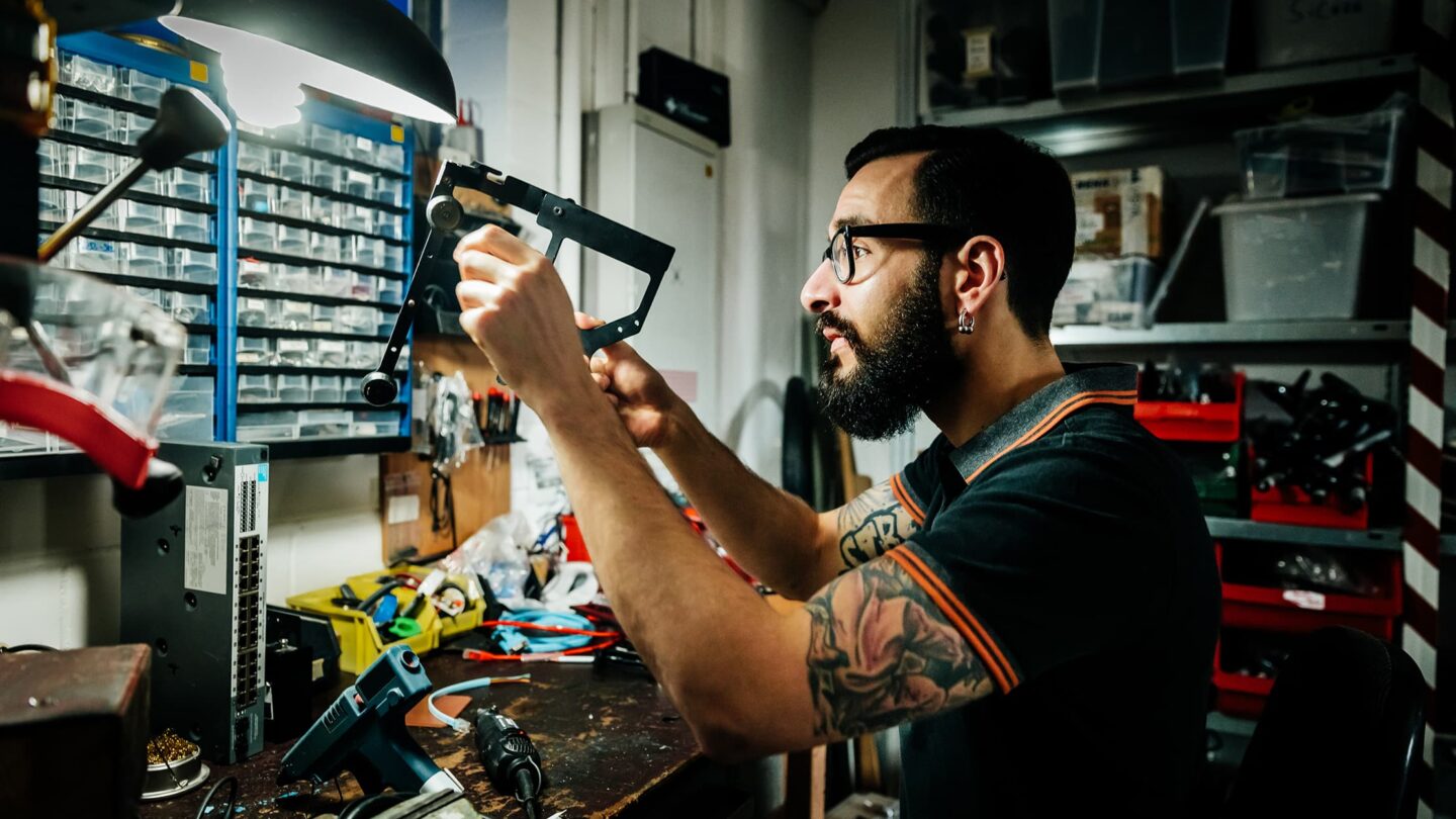 Jovem com barba a realizar obras de renovação num espaço interior, a aplicar massa numa parede com uma espátula, rodeado por ferramentas de construção e materiais de reforma.