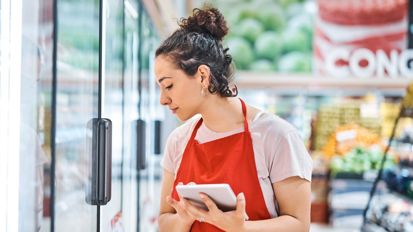 Rapariga jovem a trabalhar numa caixa de supermercado, concentrada enquanto atende um cliente. O ambiente é movimentado, com prateleiras de produtos ao fundo.