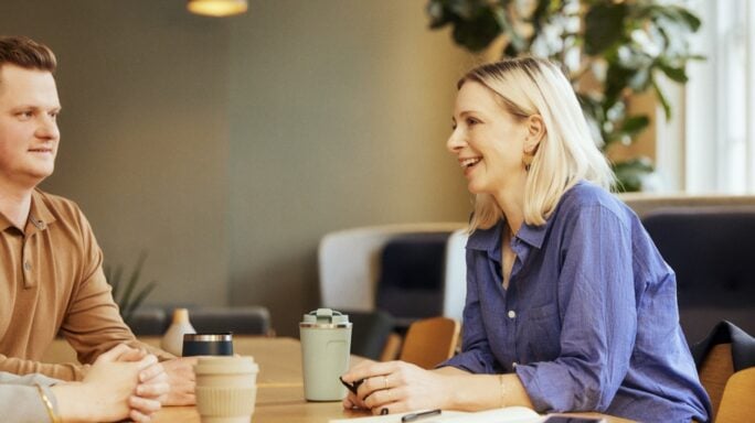 Mulher loira com camisa azul a conversar animadamente numa reunião de trabalho, enquanto os colegas, vistos de costas, escutam atentamente. Ambiente profissional e colaborativo.