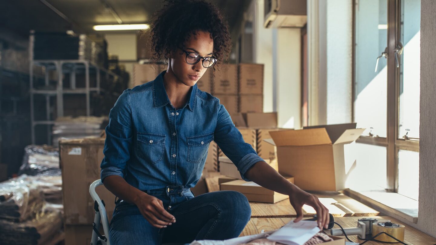 Jovem mulher sentada à mesa, concentrada no ecrã do portátil enquanto faz compras online; ao lado, um telemóvel