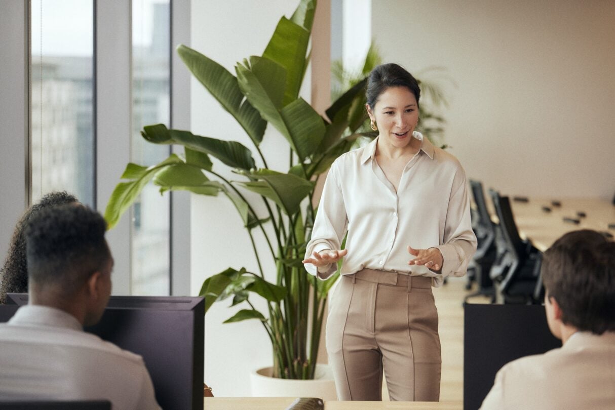 Uma mulher jovem, de cabelo castanho, a seu redor, outros colegas participam na reunião, discutindo e partilhando ideias.