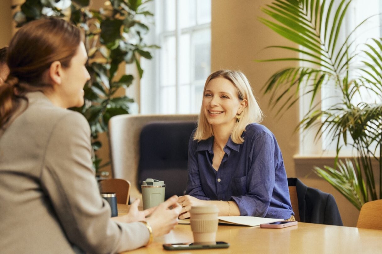 Mulher loira com camisa azul a conversar animadamente numa reunião de trabalho, enquanto os colegas, vistos de costas, escutam atentamente. Ambiente profissional e colaborativo.