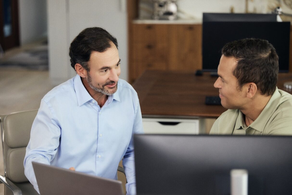 Funcionário sentado à mesa de reunião, concentrado, com documentos e portátil à frente, enquanto escuta atentamente os colegas à sua volta.